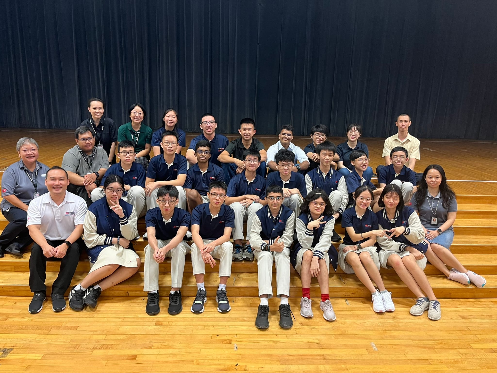 A large group of students in navy uniforms and teachers pose on wooden bleachers in front of a dark curtain.